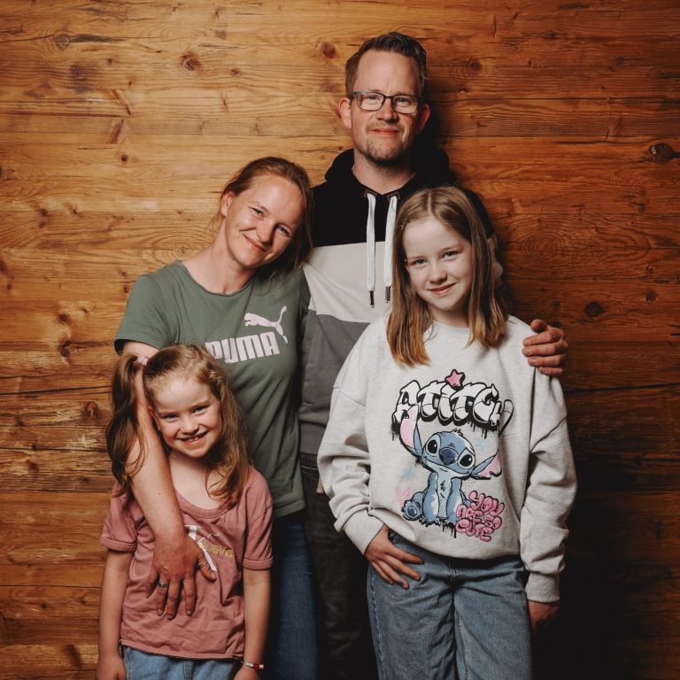 Familienfoto mit vier Personen vor einer Holzvertäfelung, lächelnd und umarmend.