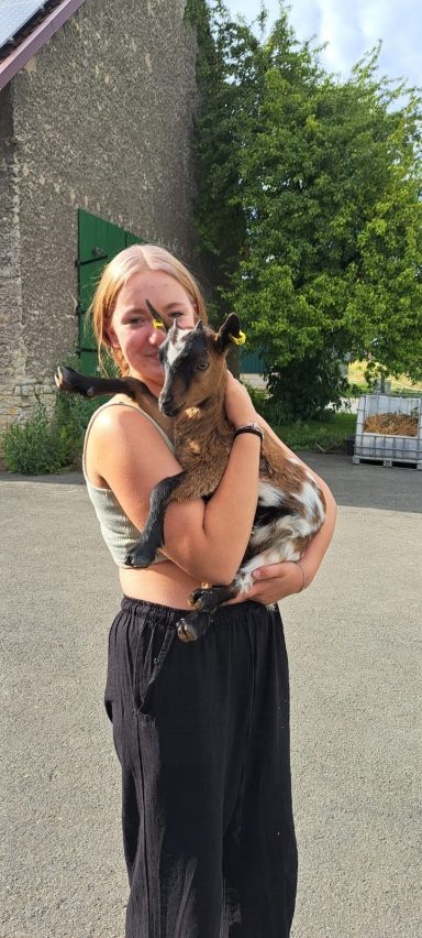 Junge Frau hält ein Zicklein vor einem Gebäude in einer ländlichen Umgebung.